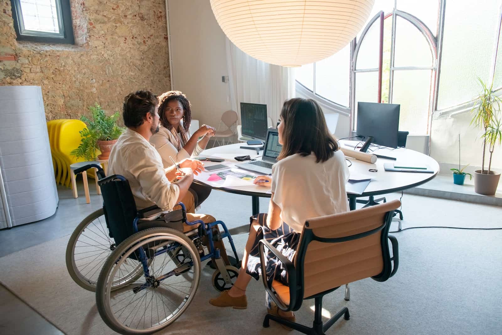 Three colleagues in a modern office setting having a discussion around a circular table, with one person in a wheelchair.