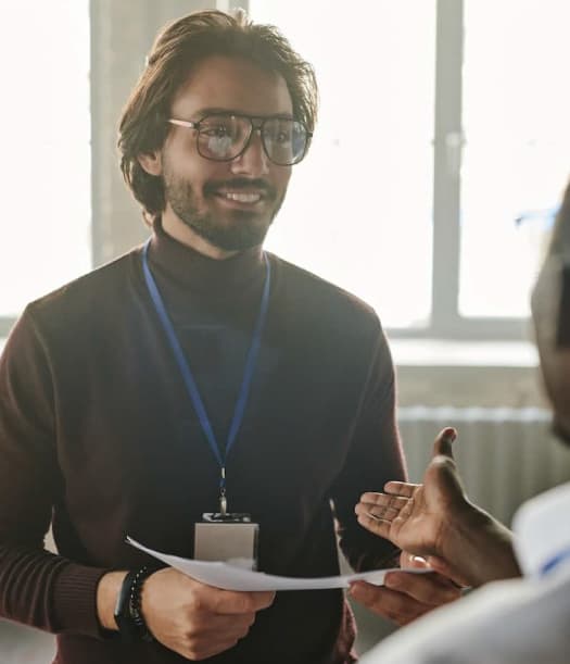 Smiling man with glasses and a lanyard engaging in a conversation with a colleague, holding a piece of paper in his hand.
